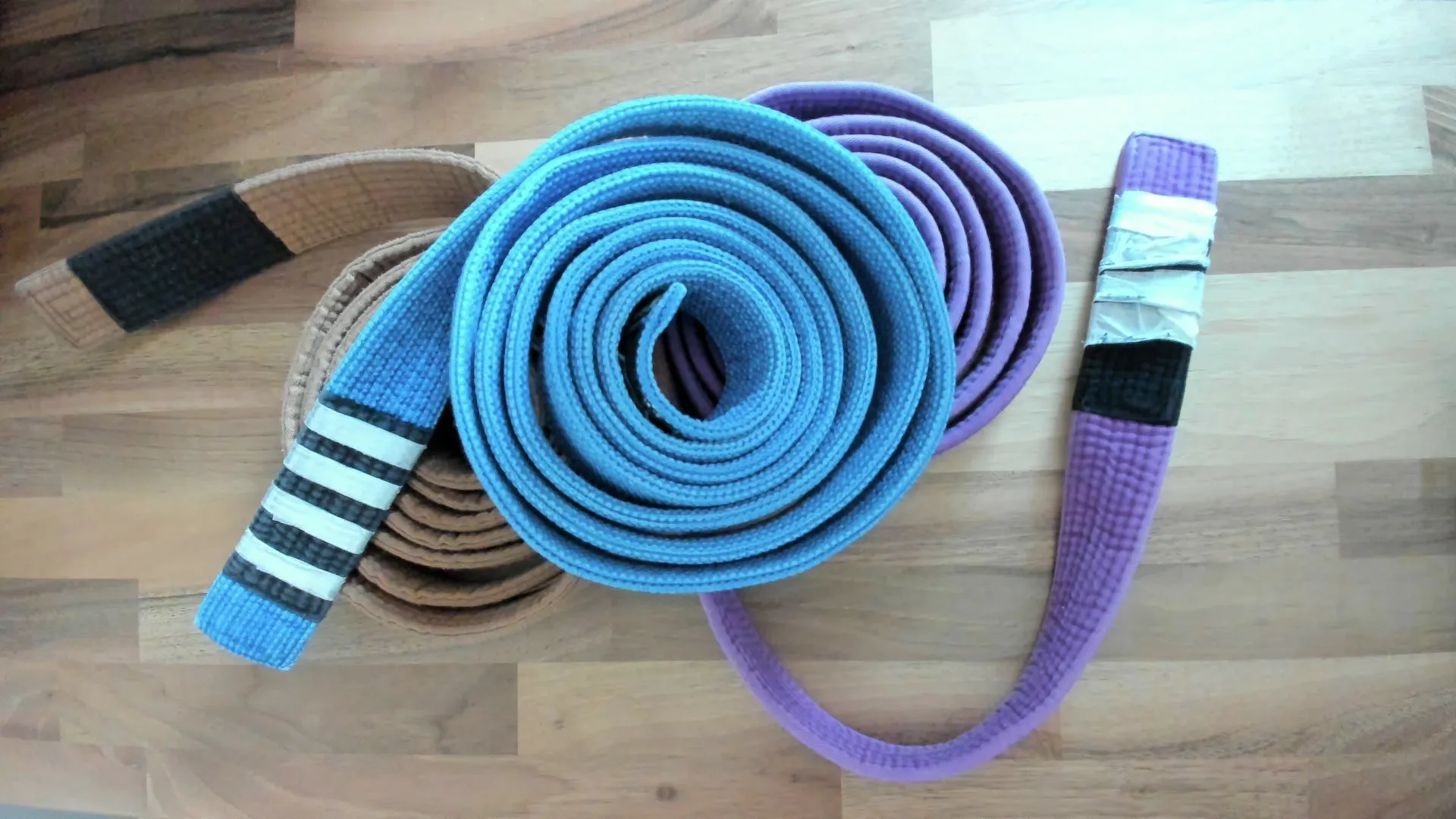Ben's BJJ belts on a wooden table, with the blue belt coiled front and centre and the purple and brown belts beside it