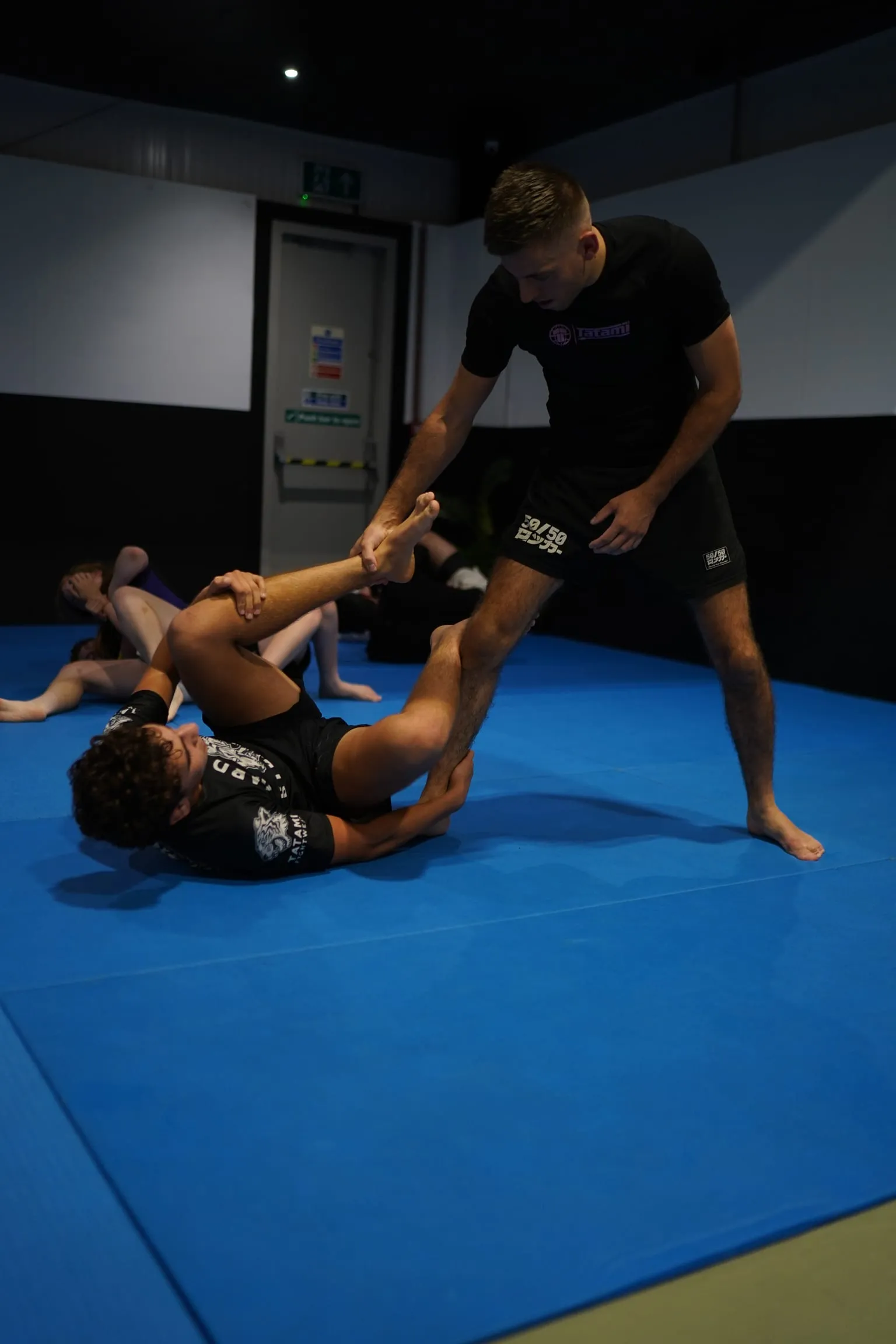 A member standing over a seated training partner and controlling one leg to pass the guard, during a no-gi class at Rebel Combat Sports