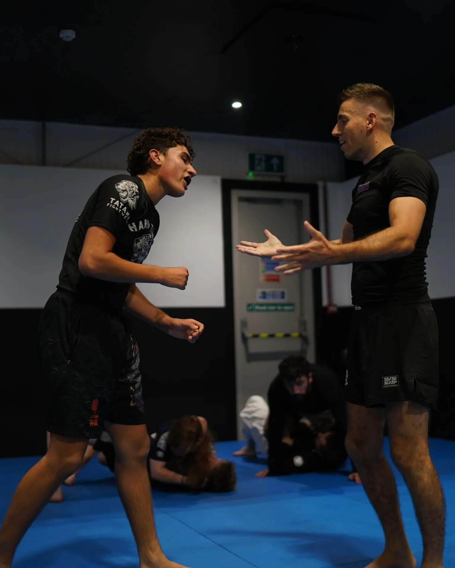 Two grapplers facing each other on their feet at the start of a round at Rebel Combat Sports, with other pairs training on the mat behind them