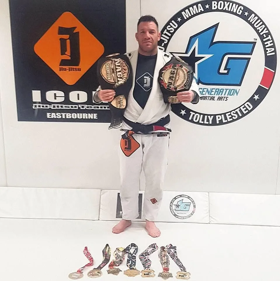 Tolly Plested in a gi and black belt at 1st Generation, holding NAGA title belts with a row of medals at his feet